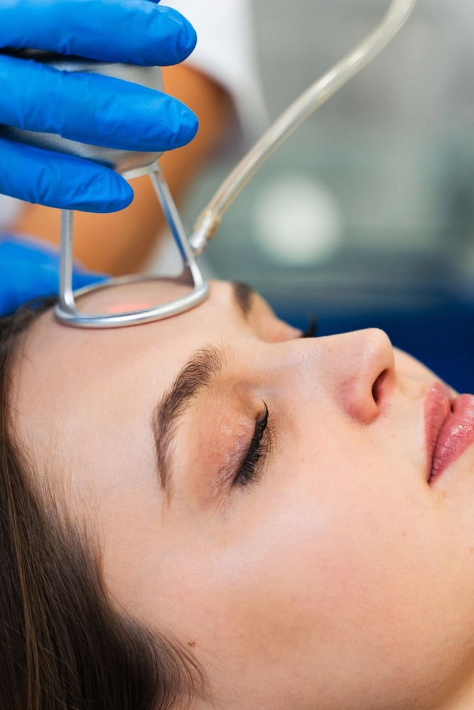 Clinician in blue gloves holding a protective eyeshield near a patient's forehead during a facial treatment