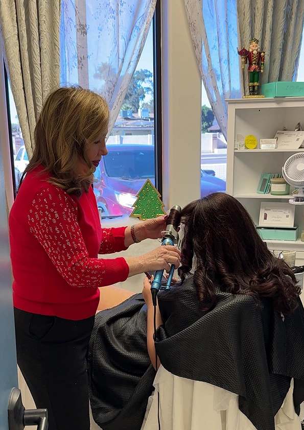 Stylist curling client's dark hair with curling iron in bright salon with bay window view
