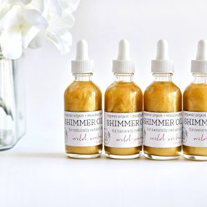 Four bottles of Shimmer Oil with organic argin and meadowfoam for naturally radiant wild orchid skin next to white flowers