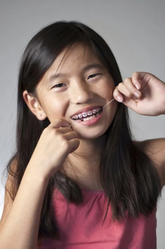 Young girl with braces flossing her teeth and smiling against a gray background