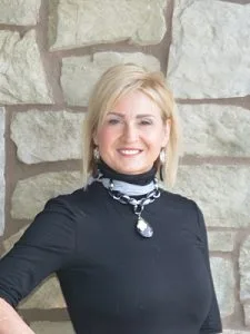 Blonde woman smiling at camera wearing black turtleneck against stone wall background