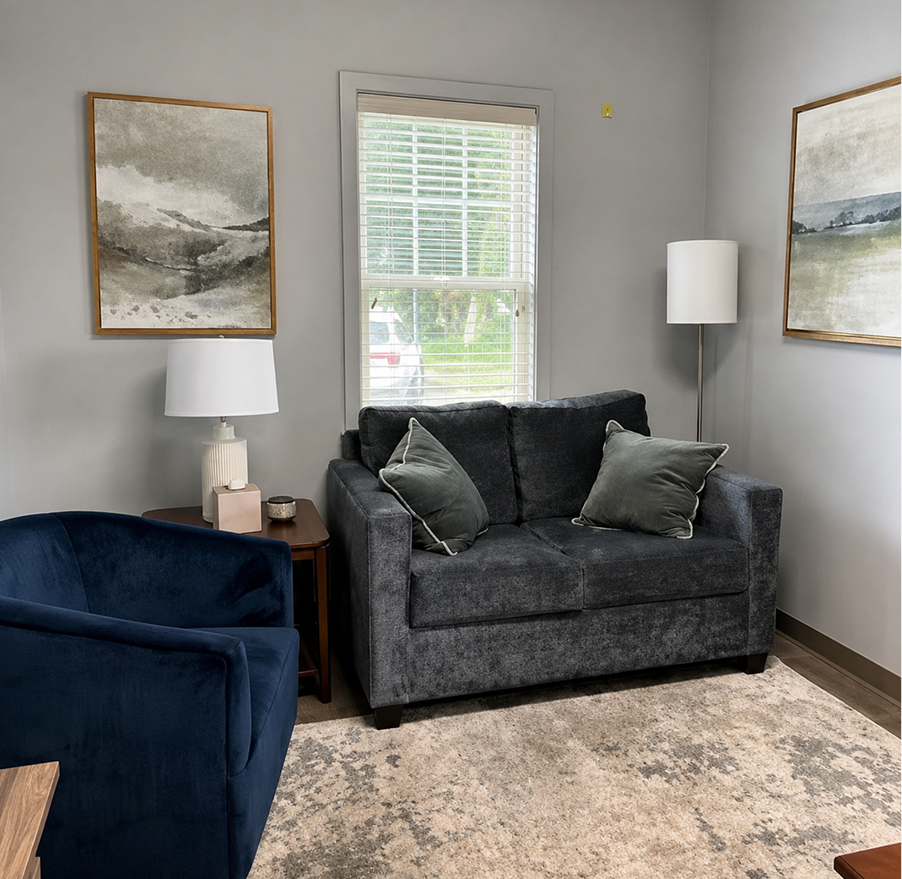 Modern office waiting area with dark gray sofa, navy armchair, framed landscape artwork, and natural lighting from large window