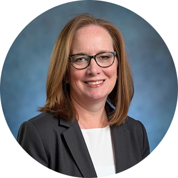 Woman with long reddish-brown hair and glasses wearing a dark blazer over white shirt, smiling at camera