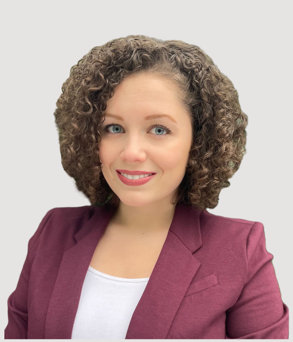 Margurite, woman with curly shoulder-length brown hair wearing a burgundy blazer, smiling at camera