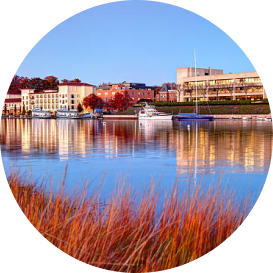 Waterfront cityscape with brick buildings reflected in calm water and marsh grasses in foreground
