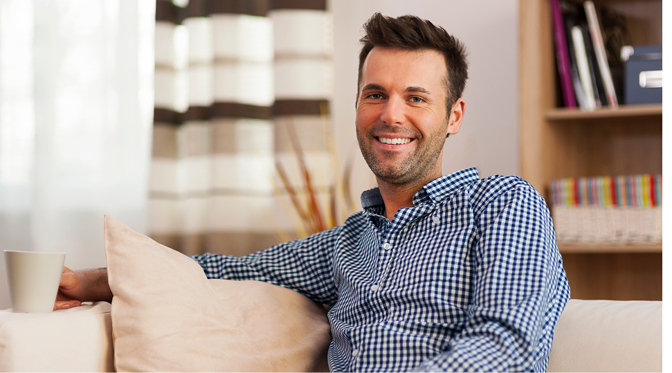 Smiling man in blue checkered shirt sitting on couch in home office with bookshelves