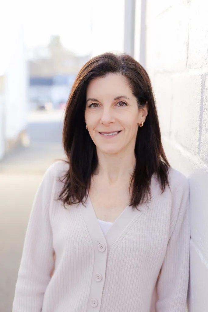 Rebecca, woman with dark hair smiling, standing by a brick wall with windows in background