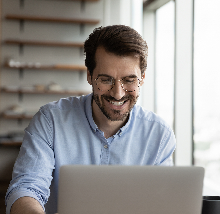 Man in blue shirt smiling while viewing laptop, sitting by window with shelving in background