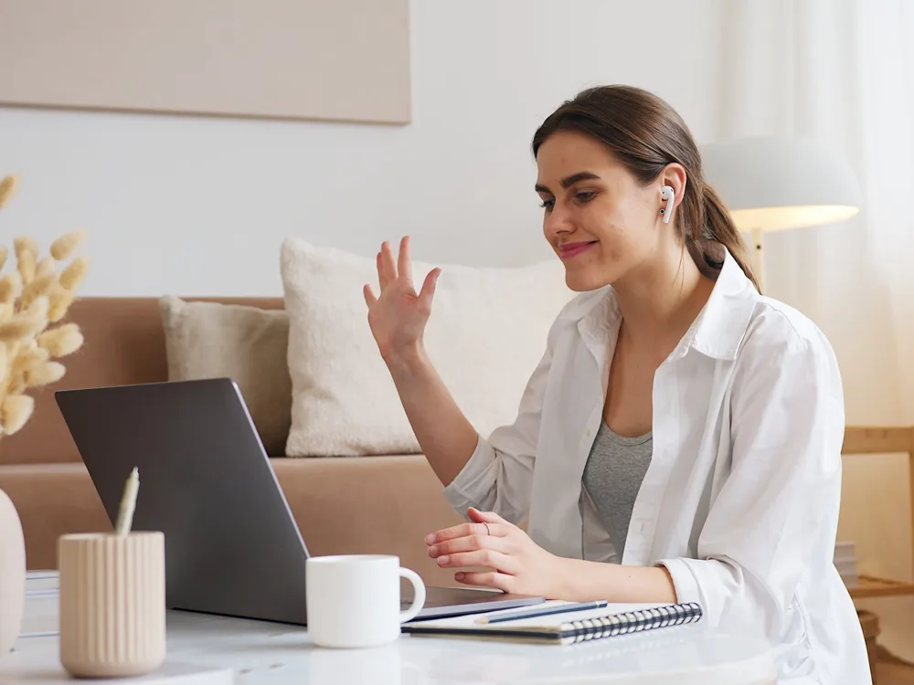 Woman with earbuds waving during a video call at home