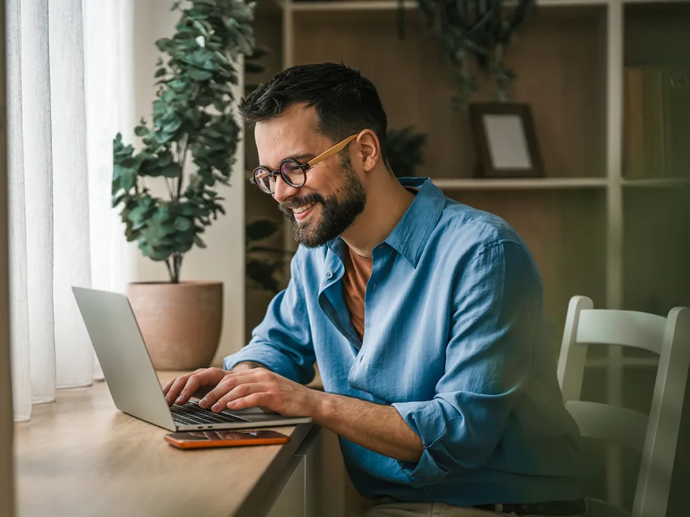 Man in glasses smiling at his laptop at a wooden desk