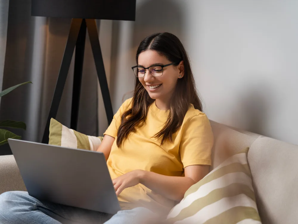 Woman in glasses smiling at her laptop on the couch