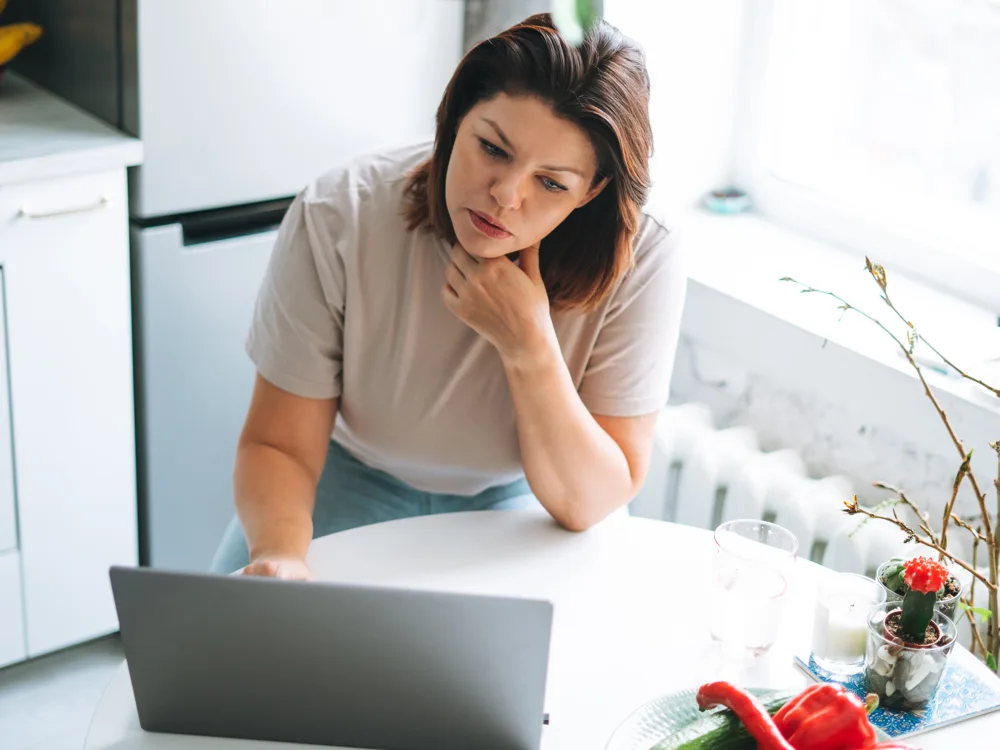 Woman leaning over a kitchen table looking at her laptop