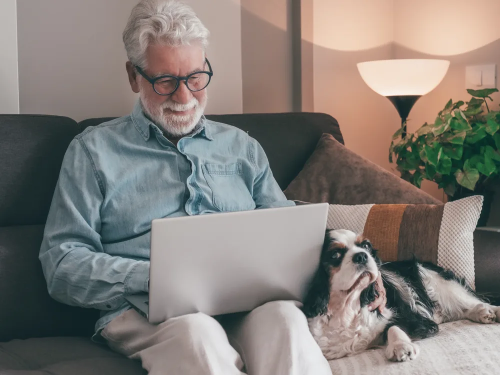Older man smiling at his laptop on the couch with his dog