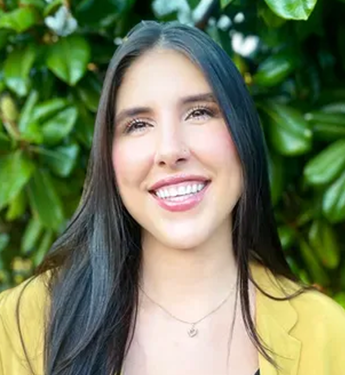 Emily Brangwin smiling at the camera, wearing a yellow top with green foliage in the background