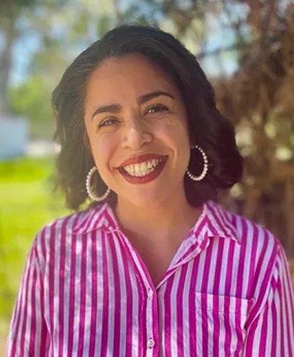 Woman smiling outdoors wearing magenta striped shirt with hoop earrings, tree in background