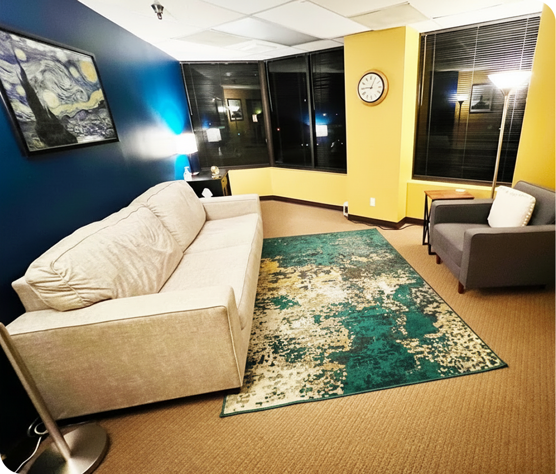 Daybreak Counseling Center Cerritos therapy room with cream sofa, gray armchair, teal and gold rug, blue and yellow walls, and framed artwork