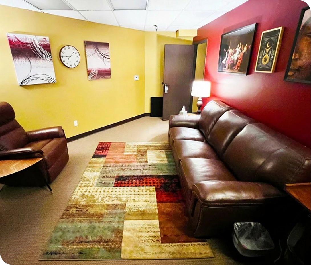 Daybreak Counseling Center Cerritos therapy room with burgundy leather sofa, brown armchair, multicolored rug, yellow and red walls with framed art and clock