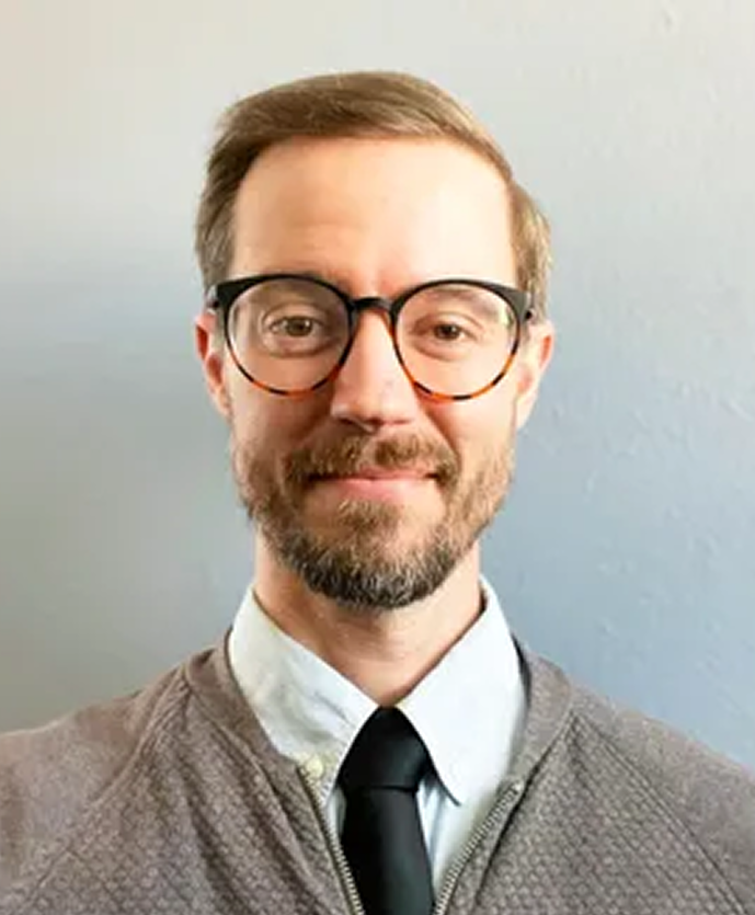 Luke wearing glasses, gray cardigan, white dress shirt, and black tie, neutral background