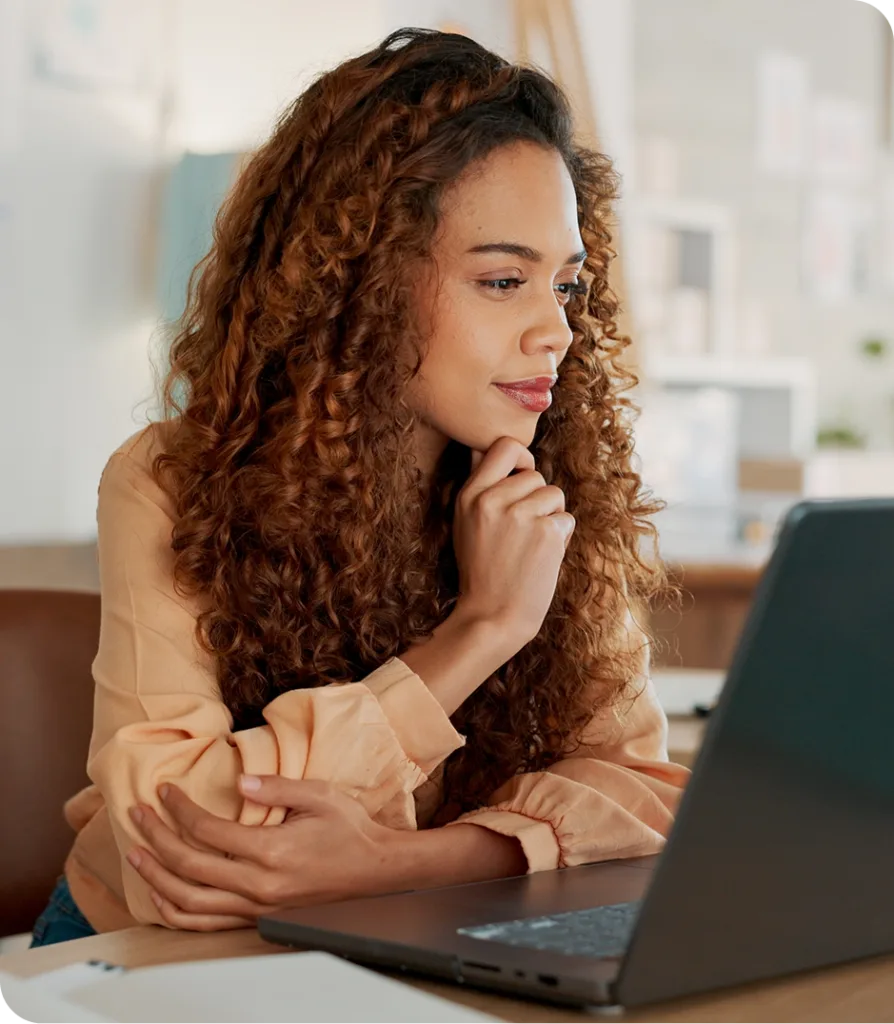 Woman with curly reddish-brown hair sitting at desk looking thoughtfully at laptop screen