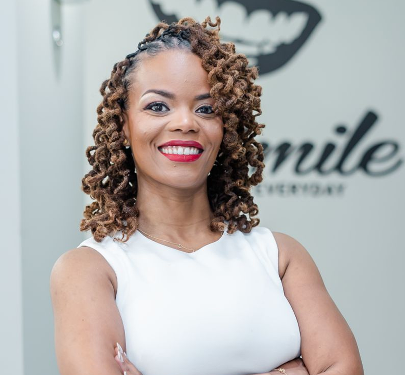 Dr. Rasheeda smiling at the camera in a white sleeveless top in front of a practice logo