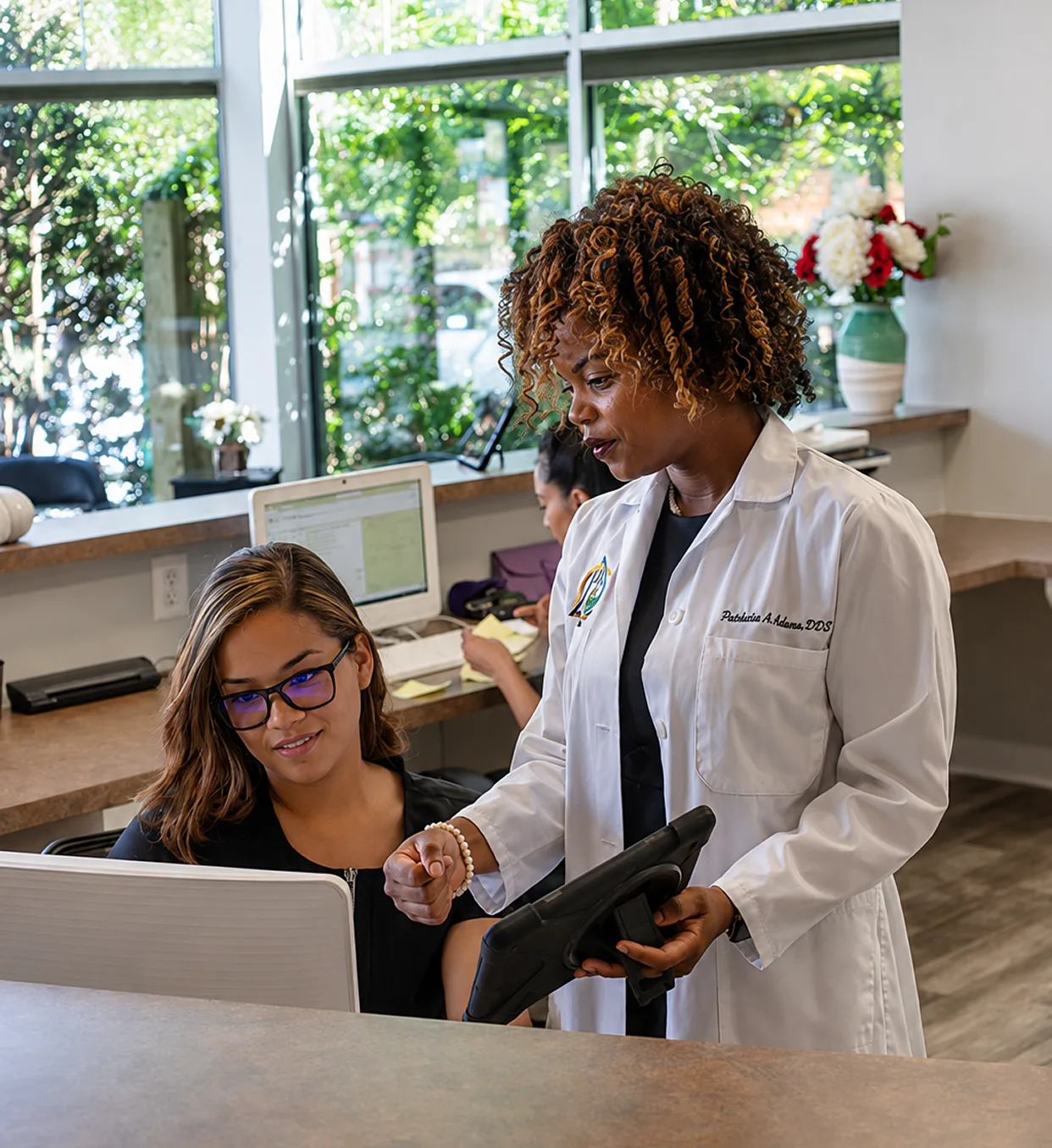Dentist in white coat reviewing tablet with patient at front desk in bright office with garden views