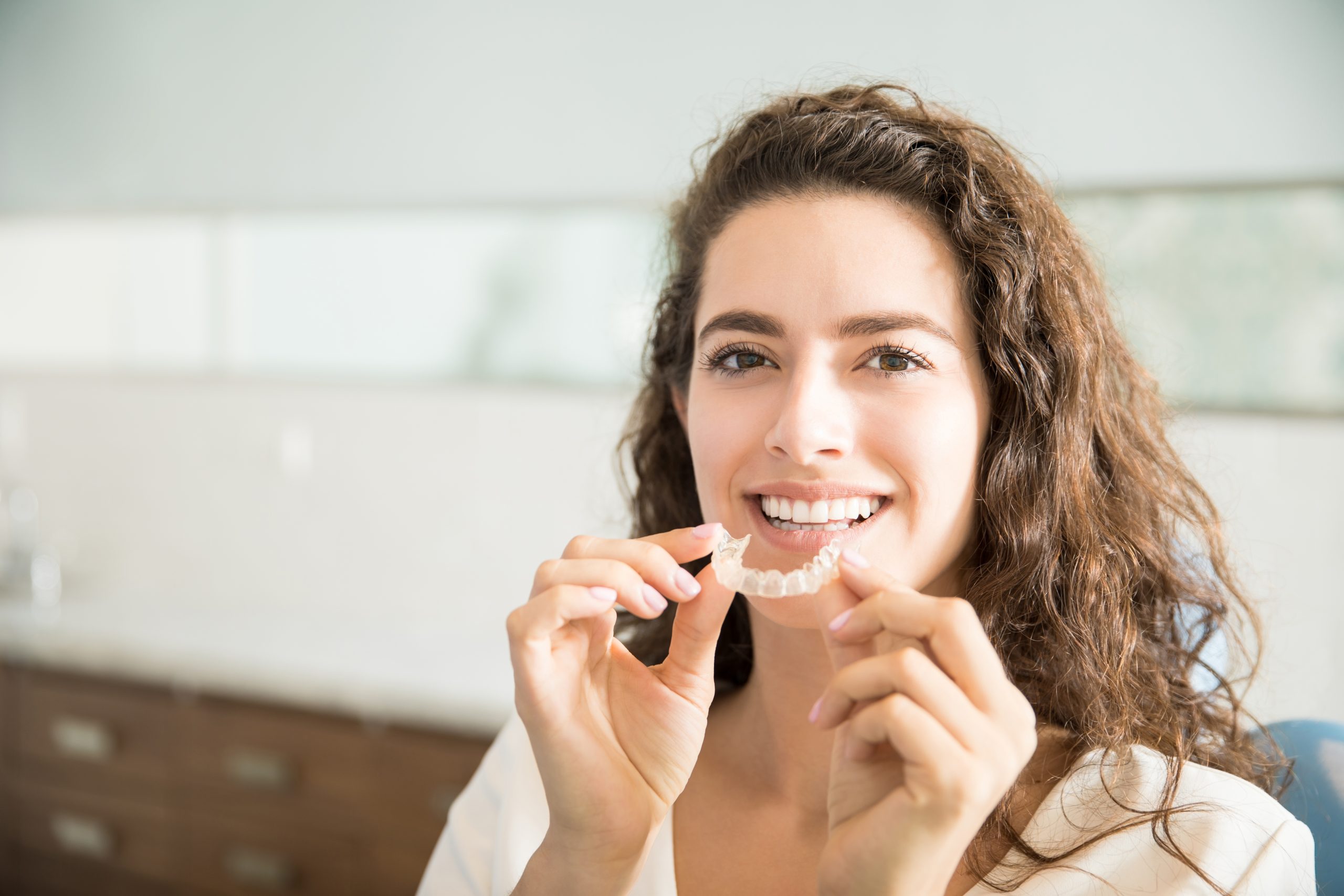 Woman smiling while holding clear orthodontic retainers in dental clinic