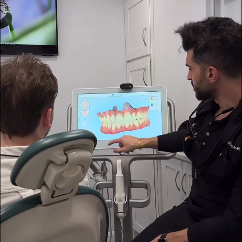 Dentist pointing to a digital 3D scan of teeth and gums on a computer monitor while consulting with a patient