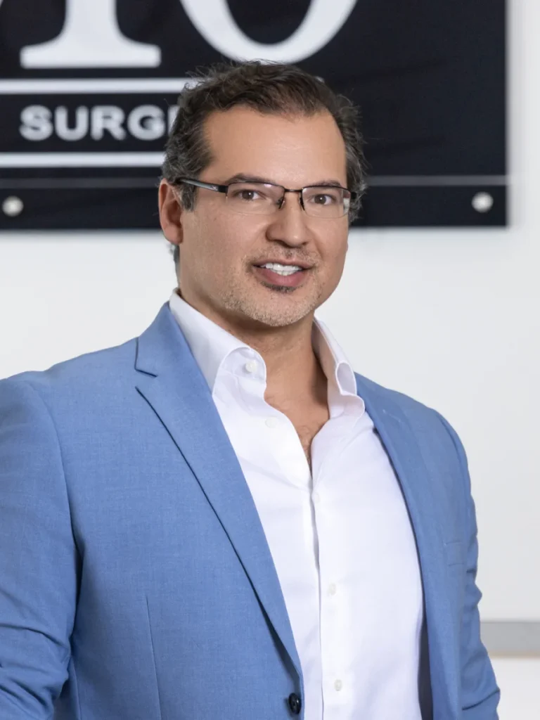 Surgeon in blue blazer and glasses smiling in front of surgical practice signage