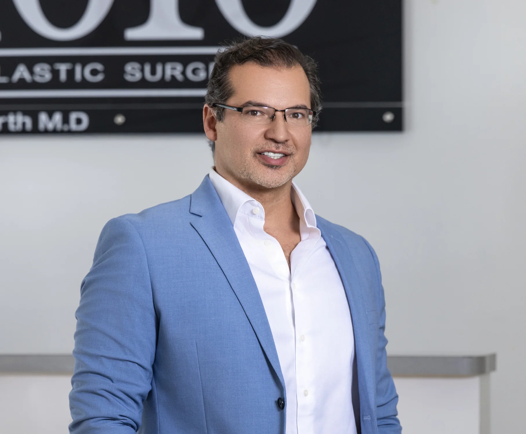 Surgeon in blue blazer and white shirt smiling in front of plastic surgery practice sign