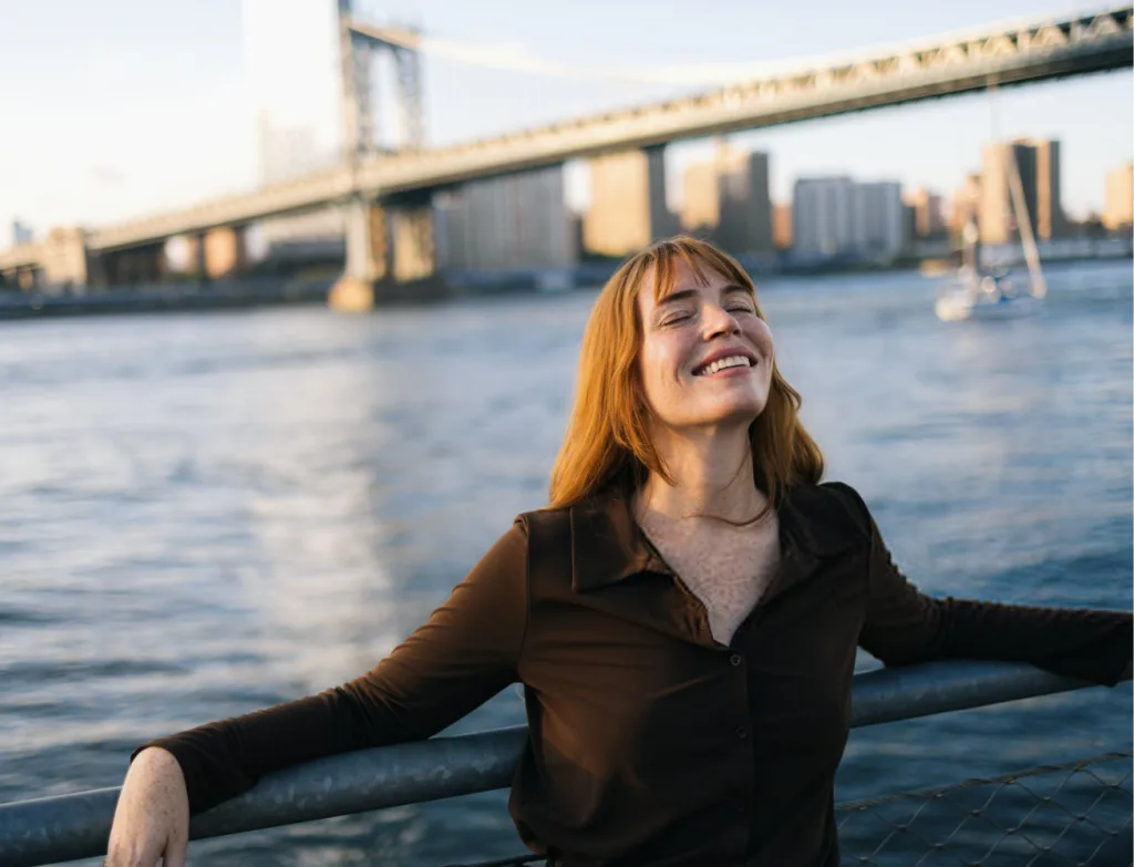 Woman with reddish-brown hair smiling at waterfront with bridge and city skyline behind her