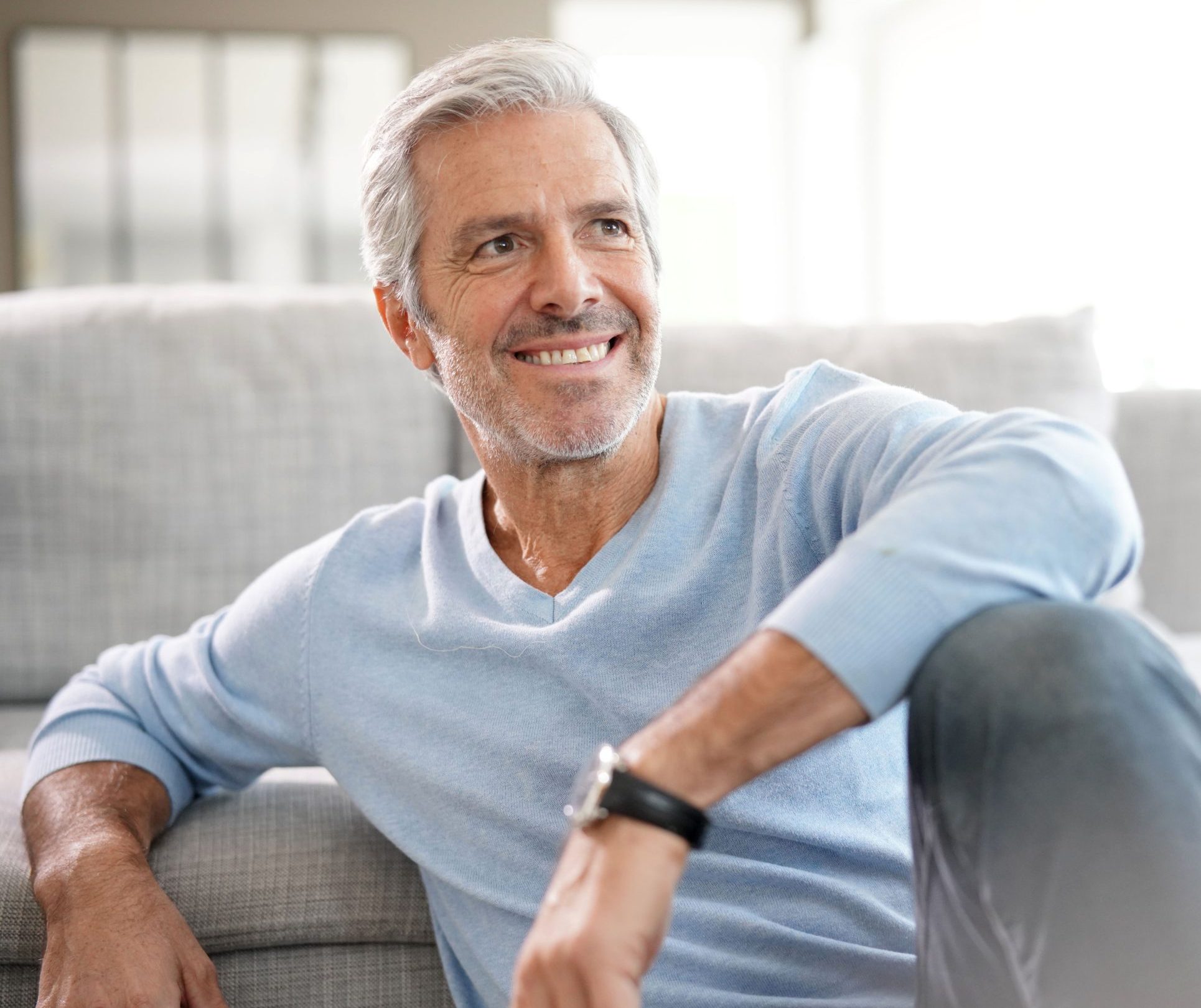 Smiling middle-aged man with gray hair sitting on a gray couch in a bright living room