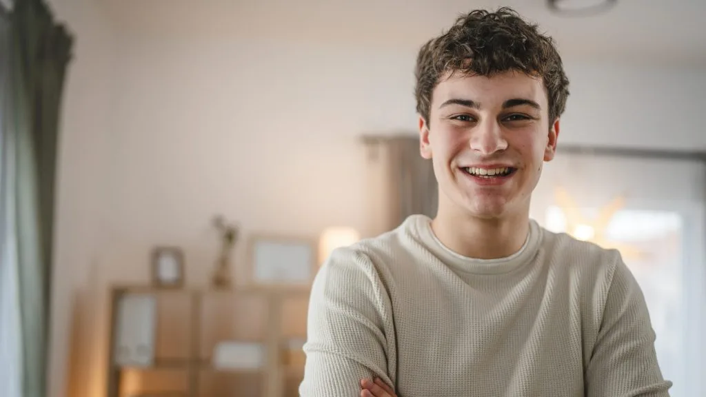 Teenage boy smiling at camera wearing a cream-colored ribbed shirt in a home interior
