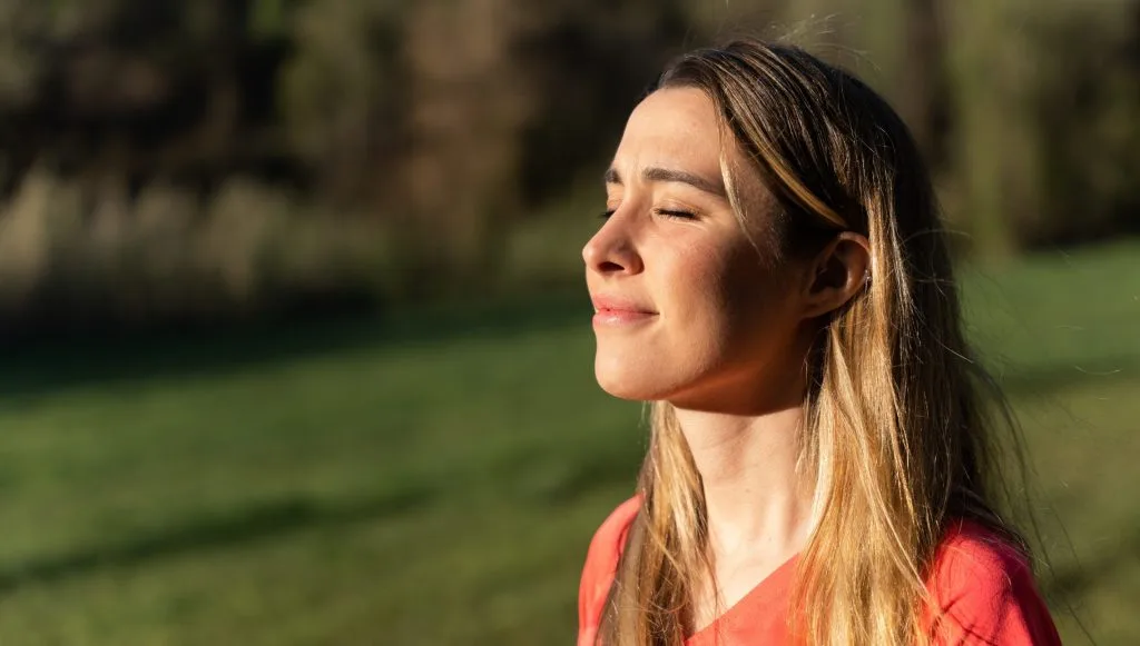 Woman with long brown hair in profile outdoors, eyes closed, wearing coral shirt