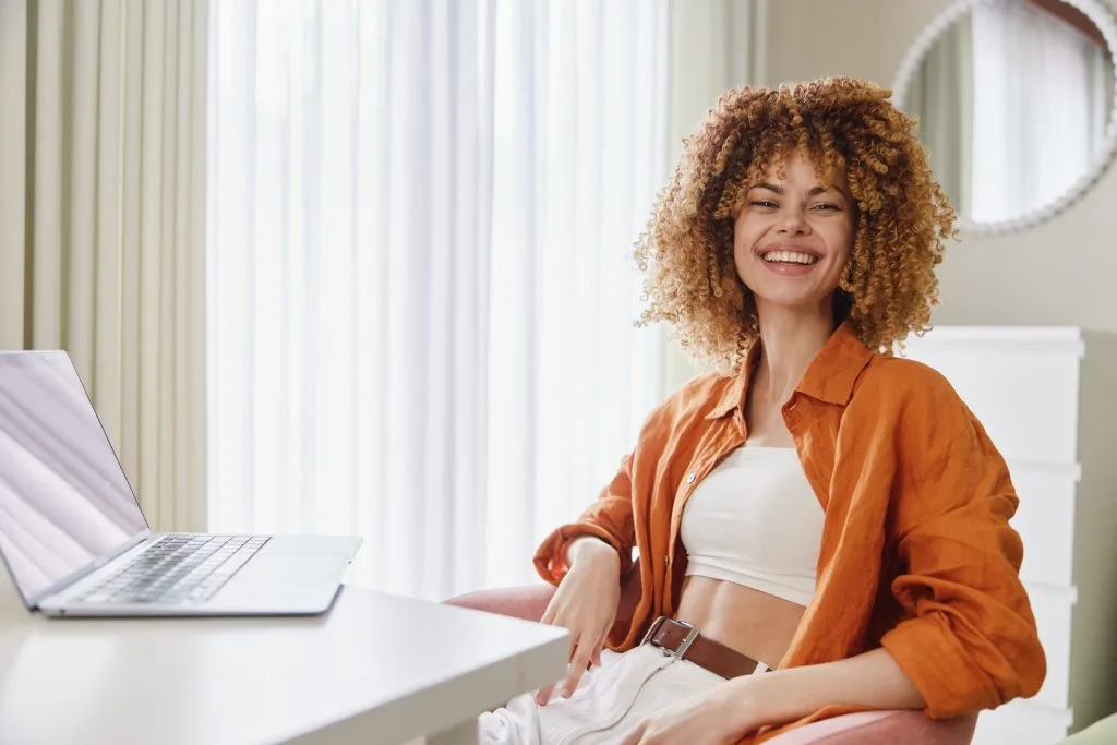 Woman in orange shirt sitting at desk with laptop, smiling indoors