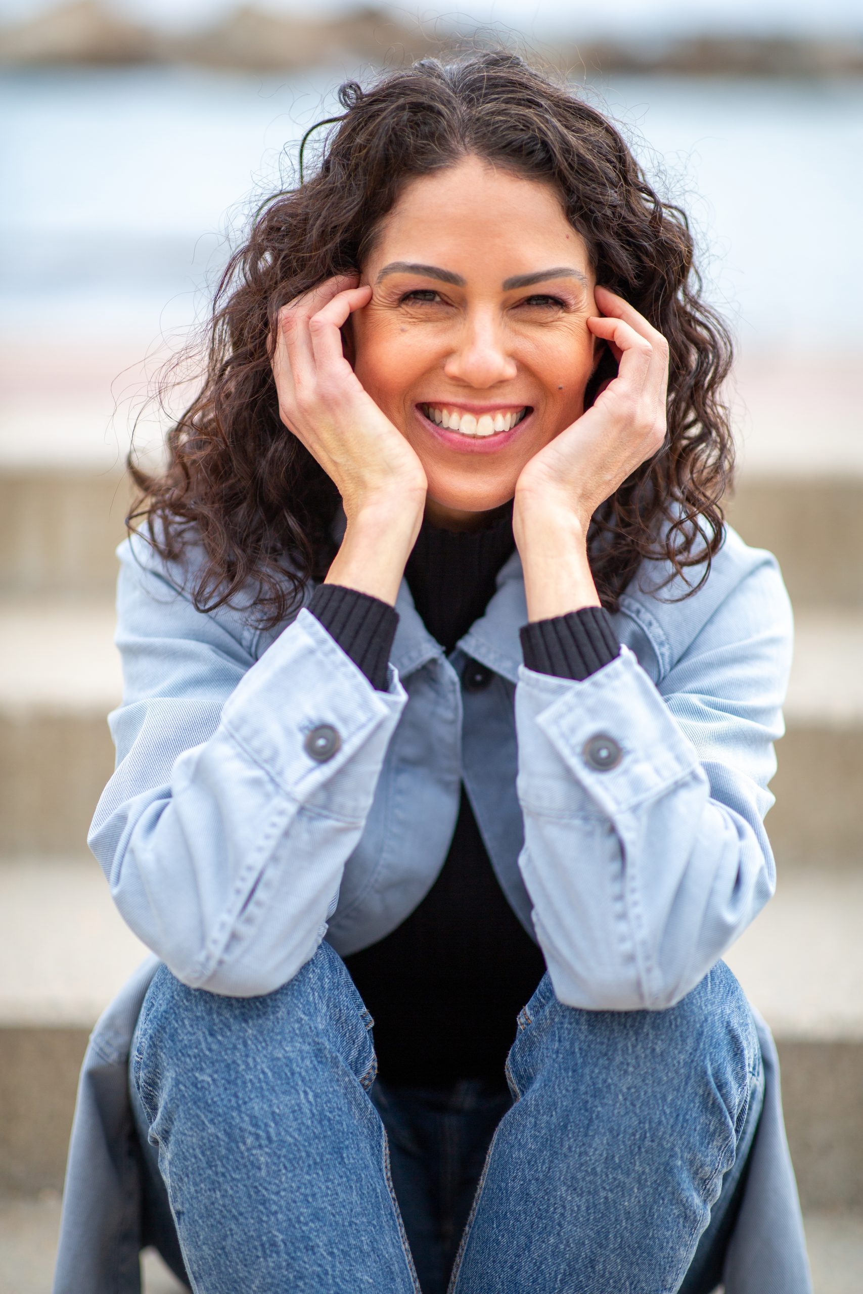 Woman with curly dark hair smiling with hands near face at beach