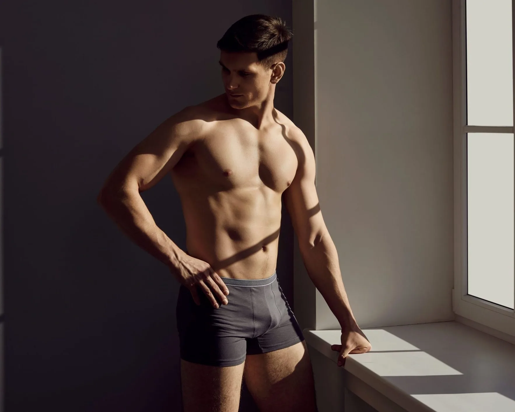 Shirtless man posing by window with hand on hip wearing gray shorts