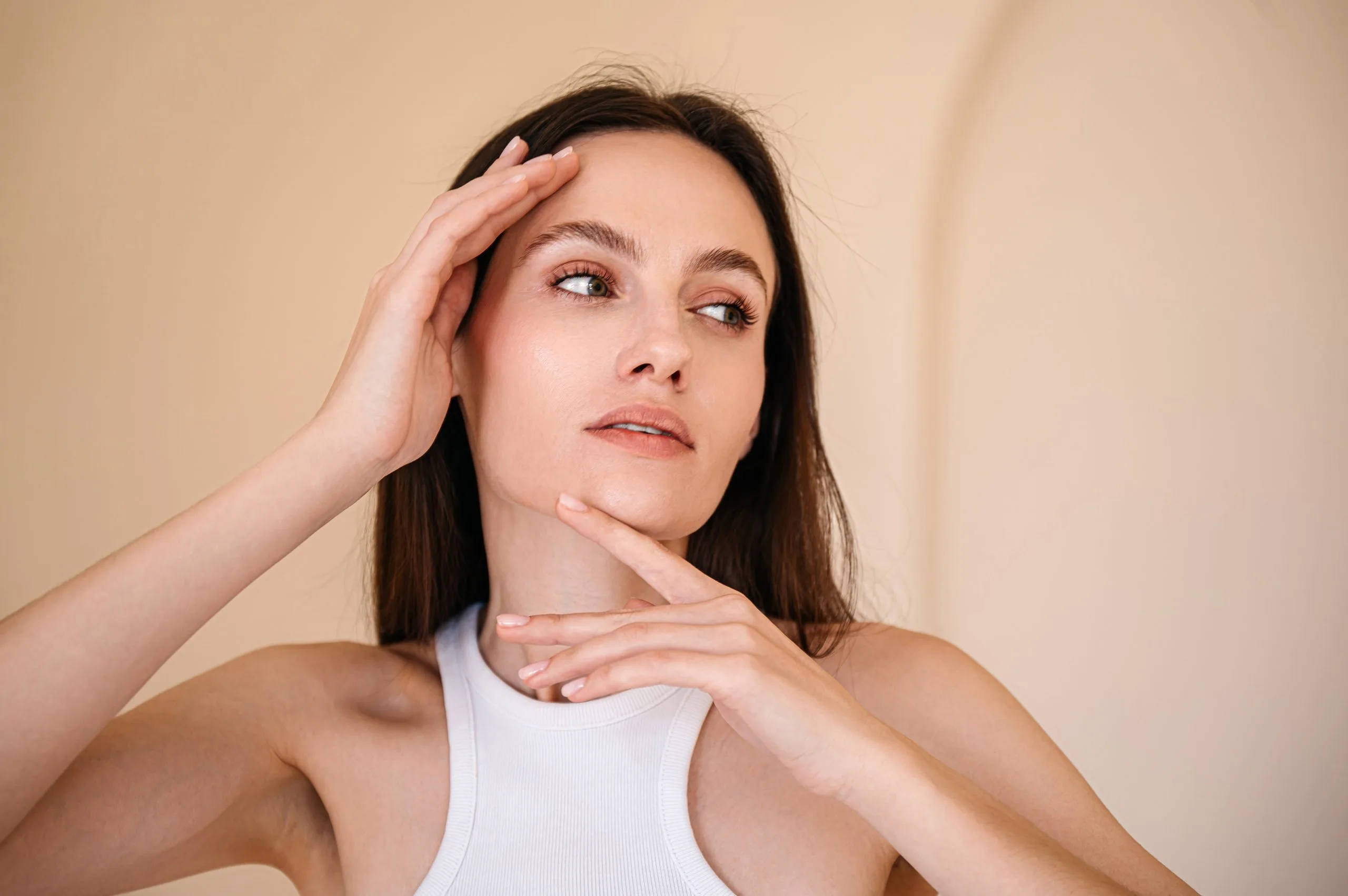 Woman with dark hair touching her face and looking thoughtful against beige background