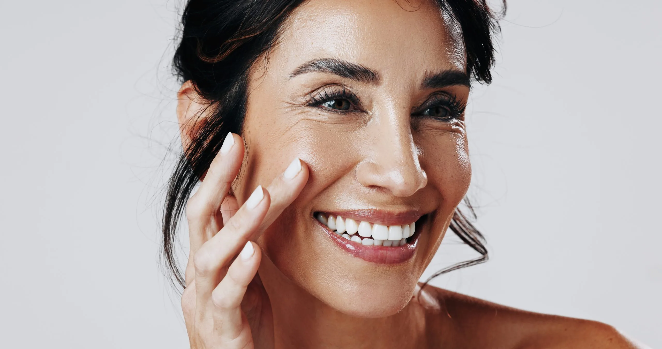 Woman with dark hair smiling broadly while touching her cheek with hand against white background