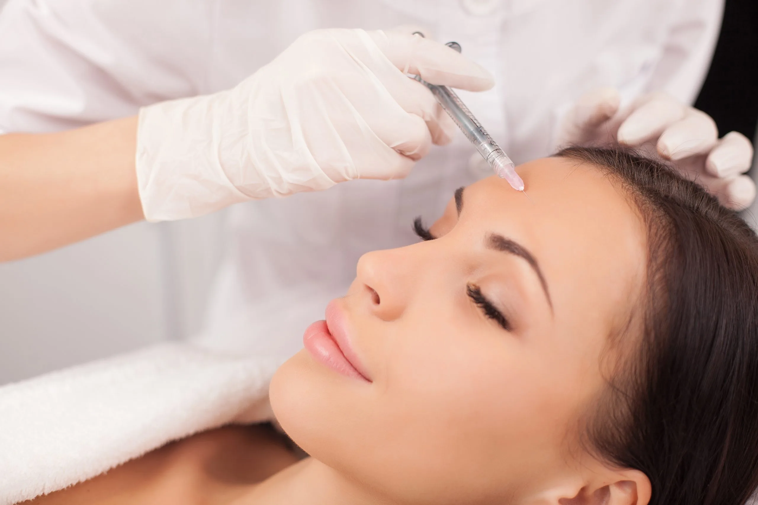 Clinician in white gloves injecting filler into patient's forehead area during cosmetic treatment