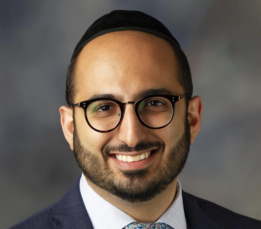 Professional headshot of smiling man wearing glasses and dark blazer
