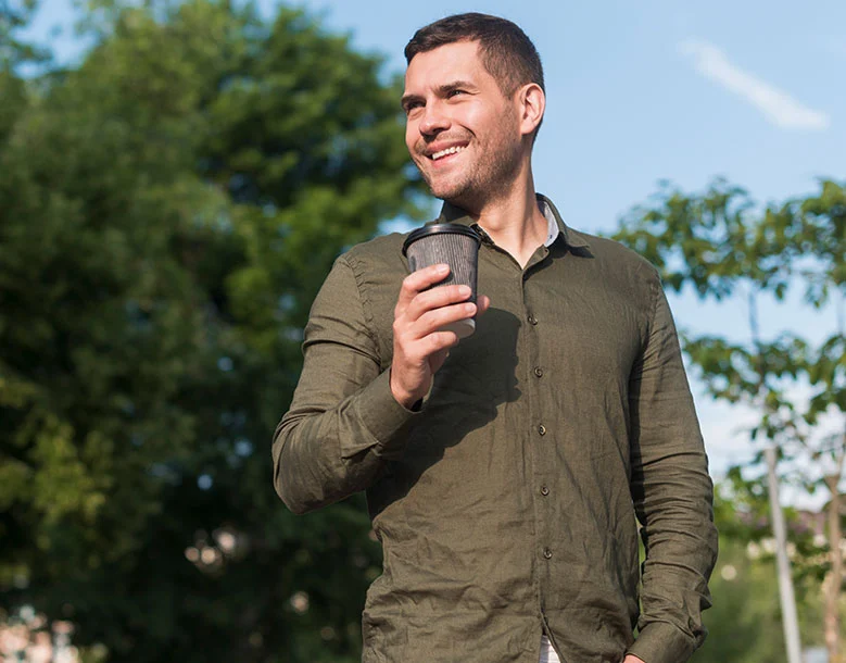 Man holding coffee cup outdoors smiling with trees and blue sky in background
