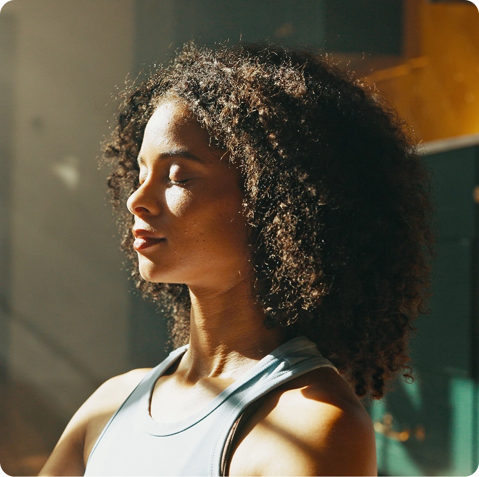 Woman with curly hair in profile, eyes closed, wearing white tank top indoors