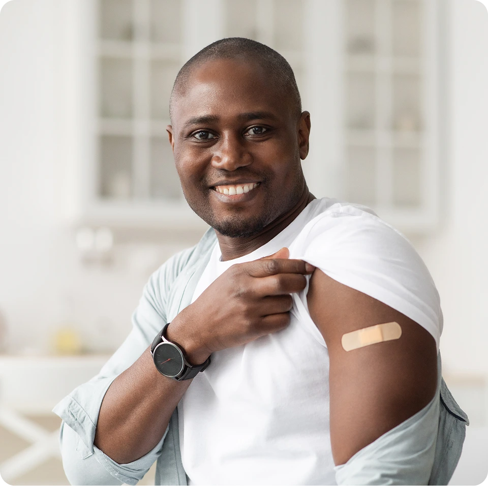 Man in white shirt pointing to bandage on his arm, smiling at camera