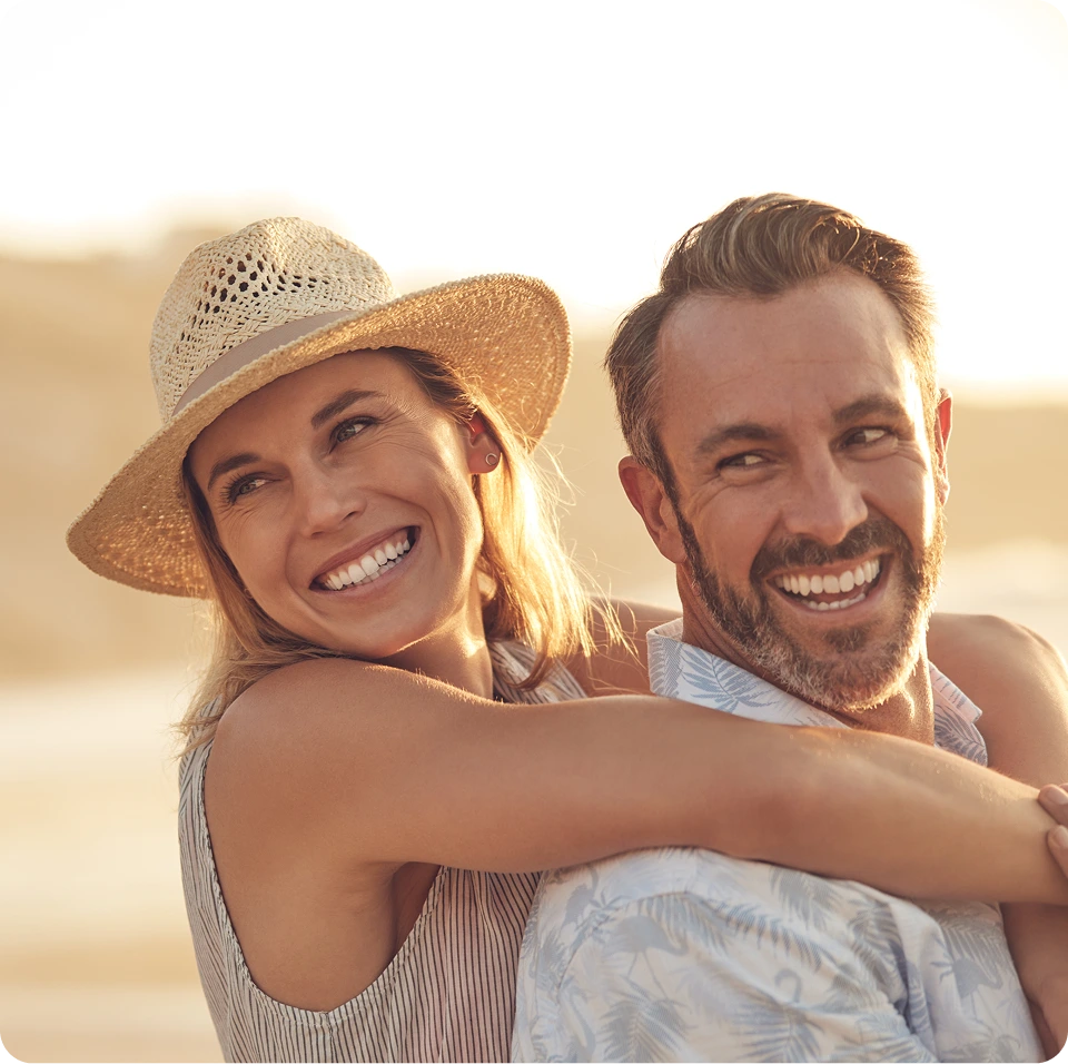 Couple smiling at beach, woman wearing straw hat with arm around man's shoulder