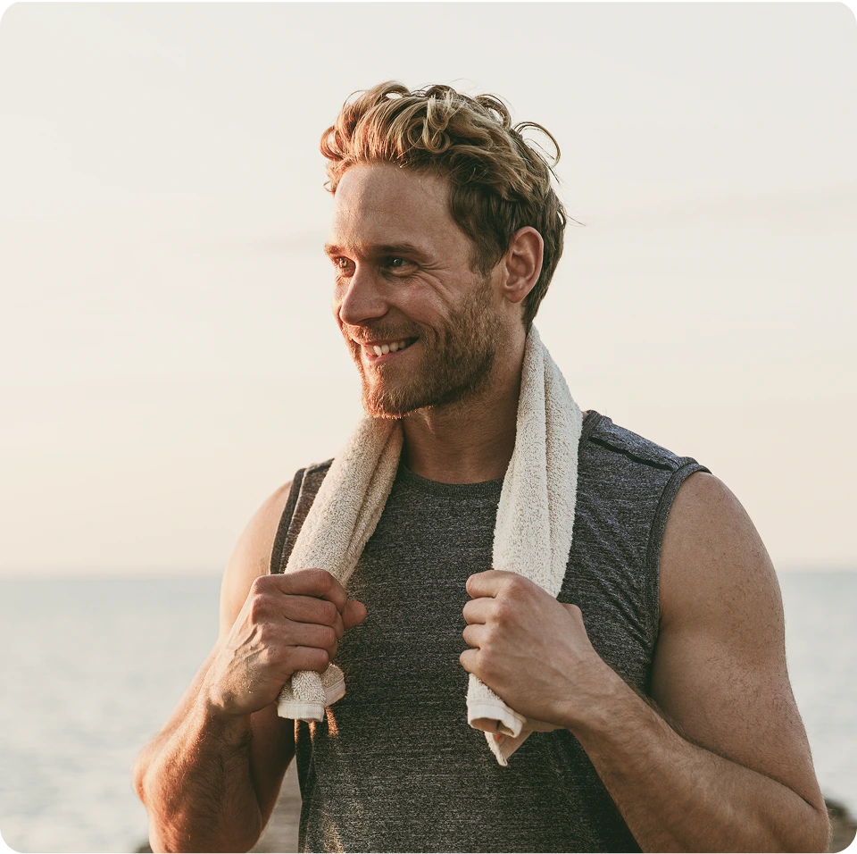 Man smiling at beach holding towel around neck, wearing sleeveless shirt