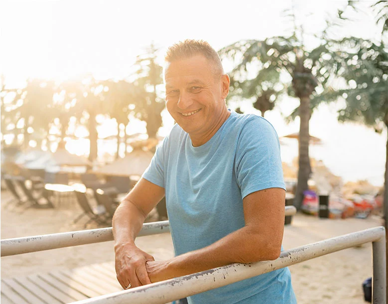 Man in blue shirt leaning on railing at beach at sunset, smiling