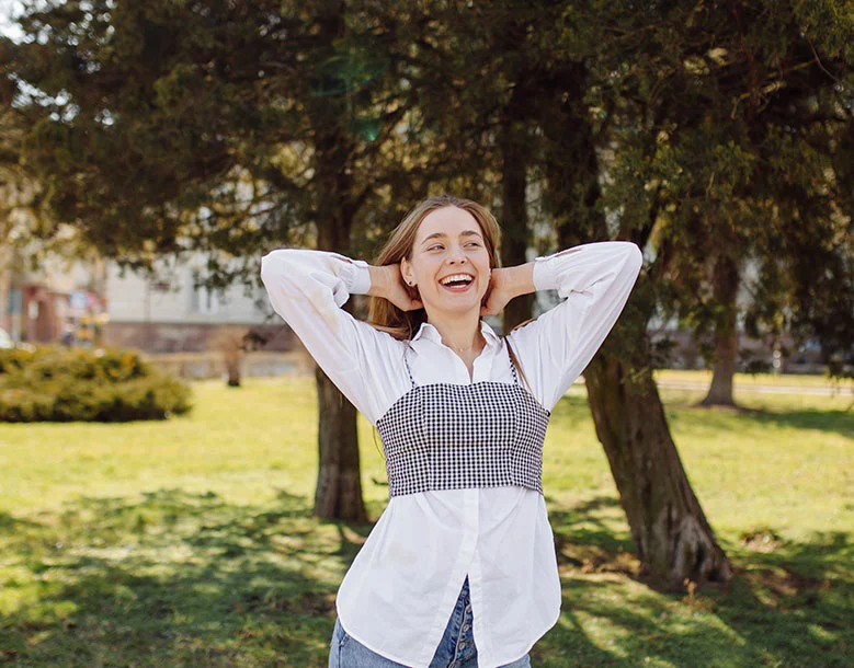 Woman smiling with hands behind head in sunny park surrounded by trees