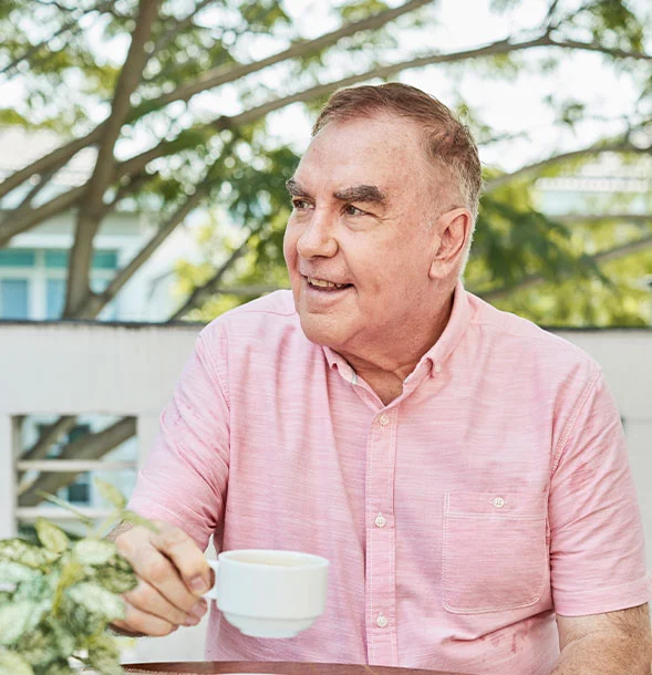 Older man in pink shirt holding coffee cup outdoors under trees