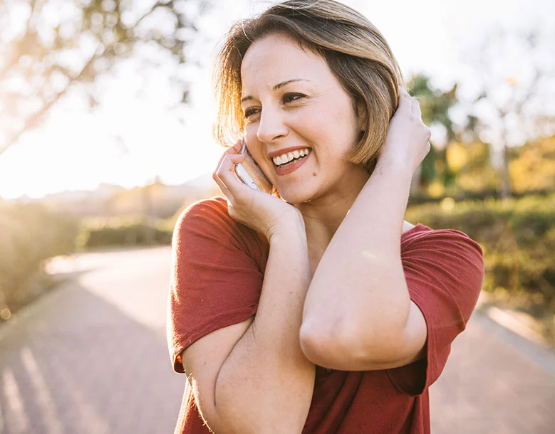 Woman smiling with hands near face outdoors on tree-lined road at sunset