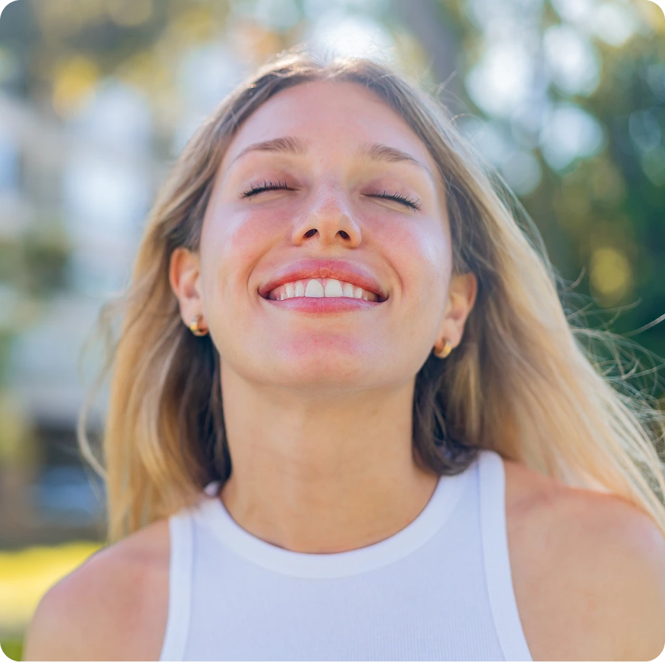 Young woman in white tank top smiling with eyes closed in natural outdoor setting
